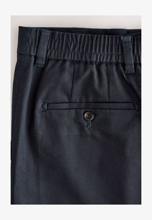 Nahaufnahme des Bundes einer dunkelblauen Hose mit elastischem Rückenteil, Gürtelschlaufen und einer einzelnen mit Knopf verschlossenen hinteren Leistentasche.