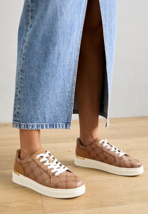 Personne portant des baskets marron à motifs avec des lacets blancs et une jupe en jean bleue fendue à l'avant, debout sur un sol en bois.