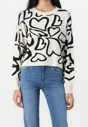 Woman wearing a white sweater with large black heart patterns and blue jeans, shown from shoulders to hips against a plain white background.