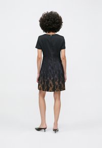 Femeie cu păr creț purtând o rochie neagră cu mâneci scurte, tiv portocaliu cu model și fermoar la spate, stând în fața unui fundal alb.