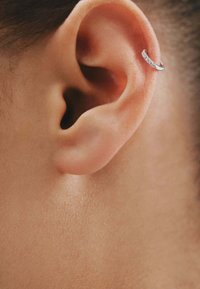 Pendiente de aro de plata con una fila de pequeños cristales transparentes. La oreja está en primer plano, mostrando el tono de piel y la textura suave.