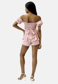 Rosa schulterfreie Playsuit mit gerafftem Oberteil, ausgestellten Ärmeln, Rüschensaum und einer Schleifenakzent an der Taille, kombiniert mit weißen High Heels.