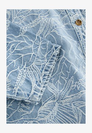 Light blue denim fabric featuring white embroidered leaf and fern patterns, with visible stitching and a brown button detail.