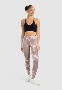 Zwarte sportbeha met geplooide details, gecombineerd met high-waisted, lichtbruine tie-dye legging. Witte sportschoenen maken de outfit compleet.