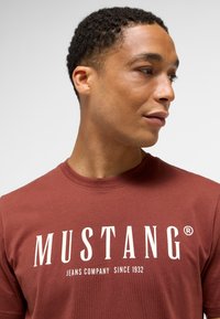 Hnědý bavlněný tričko s kulatým výstřihem, s bílým potiskem loga a textem "MUSTANG JEANS COMPANY OD ROKU 1932." Hladká textura.