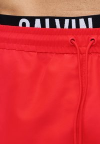 Prim-plan al zonei taliei, arătând pantaloni scurți de înot roșii cu șnururi și banda elastică neagră a chiloților Calvin Klein vizibilă deasupra.