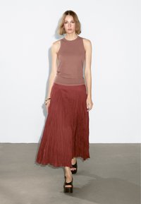 Top senza maniche color ruggine e una lunga gonna maxi plissettata. Tessuto liscio con una vestibilità morbida. La modella indossa sandali neri con tacco.