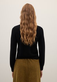 Schwarzer Pullover mit langen Ärmeln und geripptem Saum; getragen zu einem strukturierten, senffarbenen Rock mit glattem, figurbetontem Schnitt.