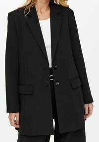 Nainen, jolla on yllään musta bleiseri, jossa on kaksi nappia ja läppätaskut, valkoisen paidan ja mustien housujen päällä, seisoo tasaisen taustan edessä.