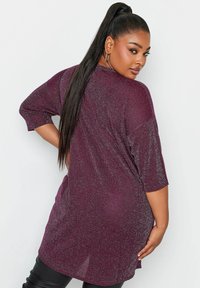 Bordowy, błyszczący oversize tunika z krótkimi rękawami, luźnym krojem i dłuższym tylnym dołem, zestawiona z czarnymi dopasowanymi spodniami.