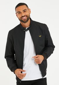 Svart bomberjacka med dragkedja, tryckknappskrage, ribbade ärmmuddar och en liten gul logotyp på bröstet. Bärs över en vit t-shirt.