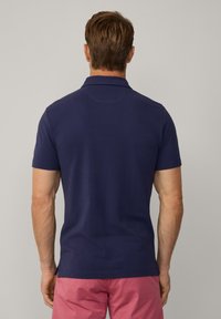 Polo shirt a maniche corte blu navy con colletto classico, texture liscia e orlo laterale spaccato, abbinato a pantaloni corti rosa, visto da dietro.