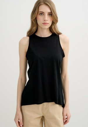 Mujer joven con cabello largo y rubio, que lleva una camiseta negra sin mangas y pantalones beige, de pie frente a un fondo claro y liso.