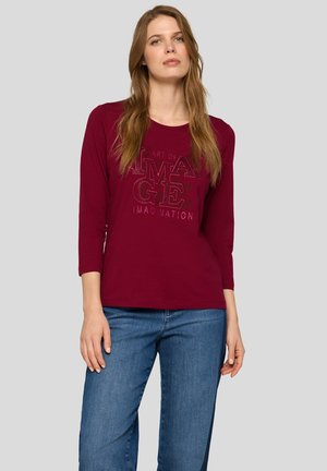Burgundy long-sleeve shirt with a round neckline featuring embossed text design. Fabric appears soft; relaxed fit. Paired with blue jeans.