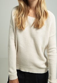 Cremfarbener gestrickter Pullover mit entspannter Passform, versehen mit einem strukturierten Muster, gerippten Bündchen und einem geraden Saum. Trägt man über schwarzen Hosen.
