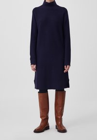 Robe en maille bleu marine avec un col montant et des manches longues, présentant des fentes latérales. Associée à des bottes hautes en cuir marron.