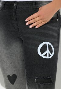 Jeans de denim negro con gráficos de símbolo de paz y corazón. Texturizados con un efecto desgastado y detalles desgastados en la rodilla.