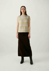 Femme aux longs cheveux bruns portant un haut sans manches beige avec des rayures roses, une longue jupe noire, et des bottines noires, se tenant devant un fond uni.