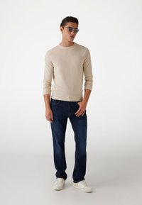 Beiger Strickpullover mit langen Ärmeln, kombiniert mit dunkelblauen Jeans und weißen Sneakers. Das Model trägt runde Sonnenbrillen.