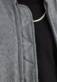Siva zipped jakna s teksturom od vune, koja ima trokutasti kviltani uzorak i logotip vez, složena preko crne majice.