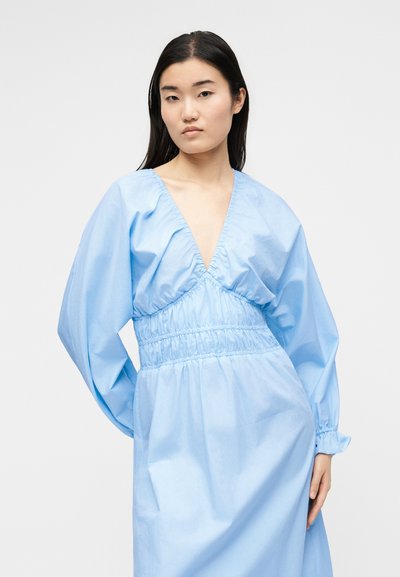 Νεαρή γυναίκα με μακριά σκούρα μαλλιά, που φοράει ανοιχτό μπλε φόρεμα με βαθιά V-λαιμόκοψη και σούρα στη μέση, στέκεται μπροστά σε απλό ανοιχτό φόντο.