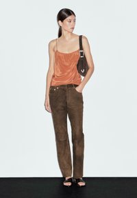 Woman wearing a copper silk camisole, brown suede pants, black sandals, and carrying a dark brown shoulder bag, standing against a white wall.