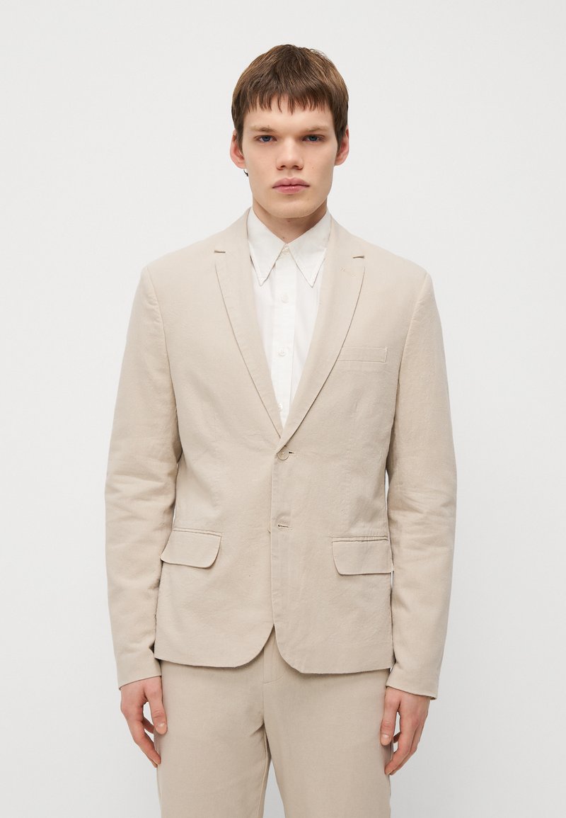 Veste de costume en lin beige avec un col cranté, deux poches avant et une texture légère. Portée sur une chemise blanche à col.