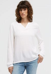 Frau mit lockigem Haar, die eine weiße Langarmbluse und hellblaue Jeans trägt und mit einer Hand in ihrer Tasche steht.