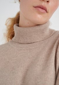 Beige turtleneck sweater made of soft knit fabric. Features a ribbed turtleneck and a textured surface, showing wrinkles and variations in color.