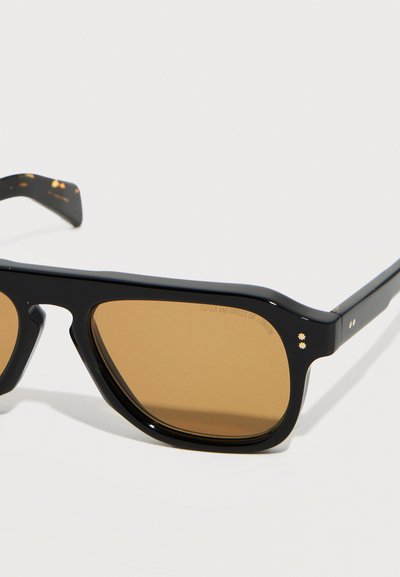 Occhiali da sole rettangolari neri con lenti marroni e piccoli dettagli in metallo a forma di stella vicino alle cerniere su sfondo bianco.