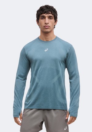 Homme portant un t-shirt de sport à manches longues bleu sarcelle et un short gris, debout face à un fond clair uni.