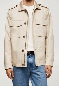 Beżowa lekka kurtka z dwoma kieszeniami na piersi, zapinana na guziki, z pagonami. Noszona na białym dzianinowym swetrze i niebieskich dżinsach.