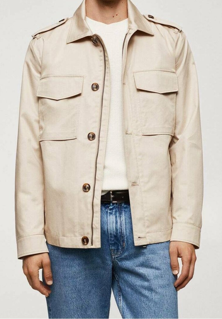 Beżowa lekka kurtka z dwoma kieszeniami na piersi, zapinana na guziki, z pagonami. Noszona na białym dzianinowym swetrze i niebieskich dżinsach.