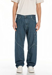 Jeans de ganga azul escuro com corte largo, detalhes de costuras angulares e bolso lateral. Usados com uma t-shirt clara e ténis brancos.