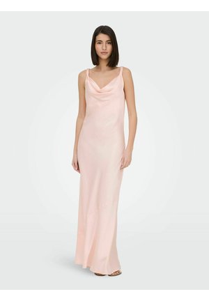 Vrouw met steil donker haar, gekleed in een lange, zachtroze satijnen nachthemd met dunne bandjes, staand tegen een effen lichte achtergrond.