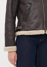 Brun skinnjakke med kremfarget skjærlingtrim på mansjettene og kanten, glidelås foran, sammen med mørkeblå denimjeans.