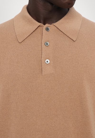 Beige kötött pólóing, amely puha tapintású, klasszikus gallérral, három gombos kivágással és finom bordázott részletekkel rendelkezik.