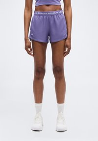 Γυναίκα με μοβ αθλητικό σορτσάκι Under Armour, λευκές κάλτσες και λευκά αθλητικά παπούτσια, στέκεται όρθια σε απλό φωτεινό φόντο.