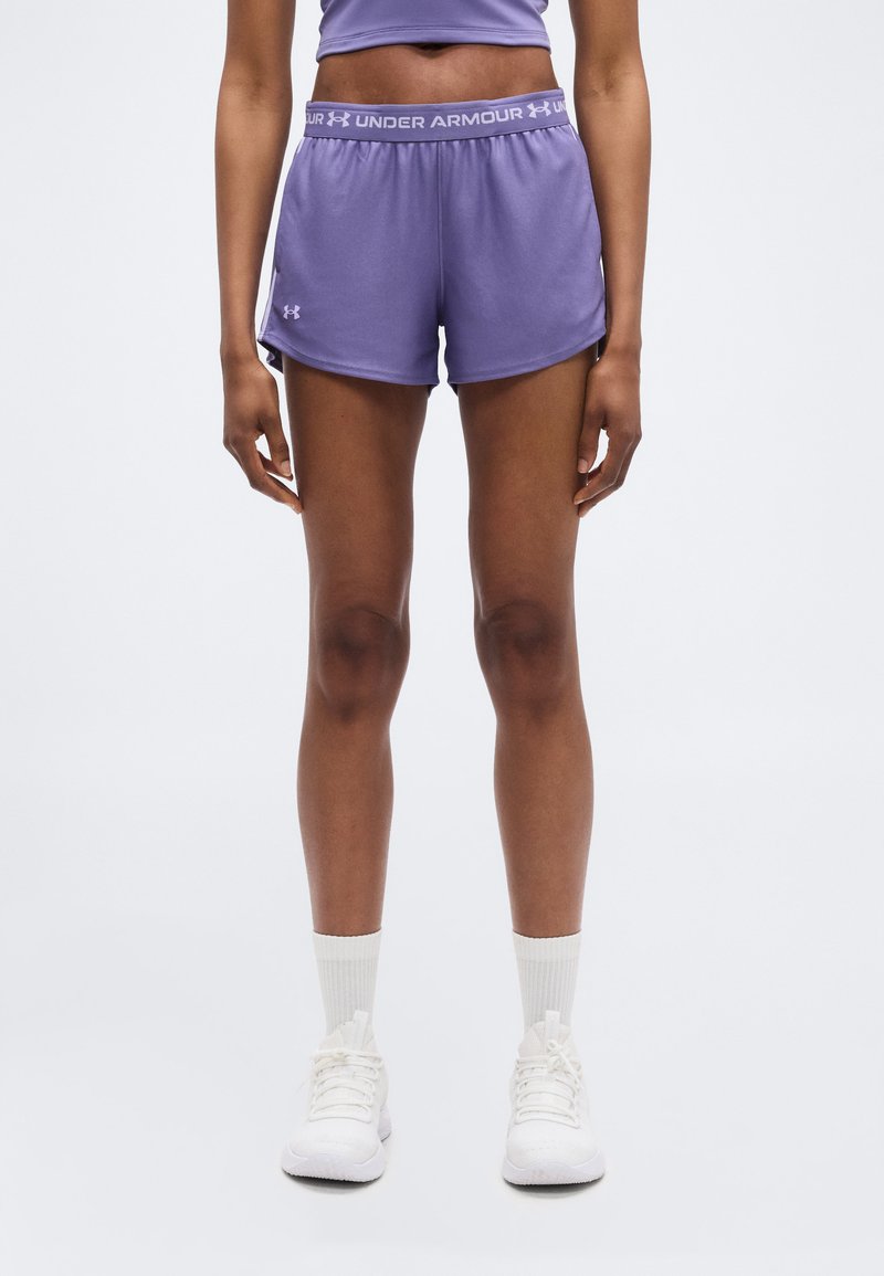 Γυναίκα με μοβ αθλητικό σορτσάκι Under Armour, λευκές κάλτσες και λευκά αθλητικά παπούτσια, στέκεται όρθια σε απλό φωτεινό φόντο.