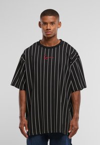 Junger Mann trägt ein übergroßes schwarzes T-Shirt mit dünnen weißen vertikalen Streifen und rotem kursivem Text auf der Brust, steht vor einem einfarbigen Hintergrund.