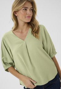 Blusa di un verde chiaro, strutturata, con scollatura a V, vestibilità ampia e maniche corte a palloncino, abbinata a denim scuro.