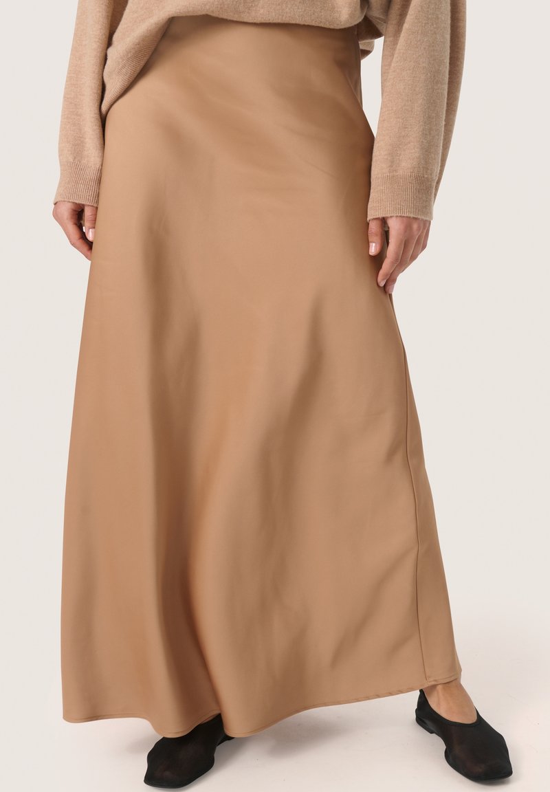 Lang, beige satin nederdel med en glat, flydende tekstur. Har en lige silhuet og en nederdel, der blødt udvider sig. Bæres med sorte flade sko.