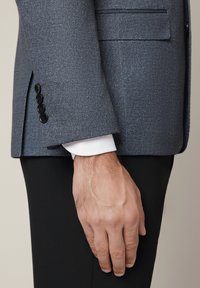 Szary, teksturowany żakiet o dopasowanym kroju, z czterema czarnymi guzikami na mankietach, noszony na białej koszuli i czarnych spodniach.