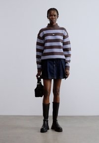 Barna és világoskék csíkos kötött pulóver, farmer szoknya, térdig érő fekete csizmák, egy kis fekete táska a kezében, egyszínű háttér.