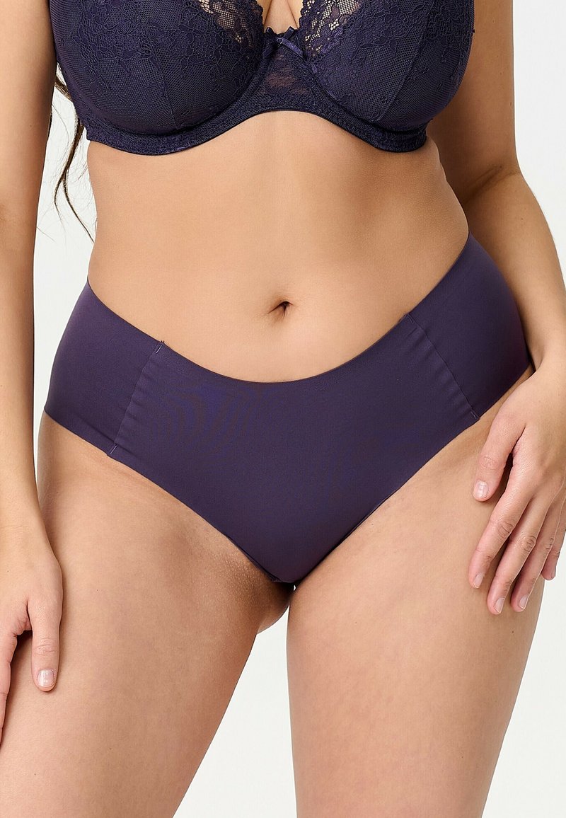 Femme portant un soutien-gorge en dentelle violet foncé et une culotte taille haute assortie, montrée du buste jusqu'aux cuisses sur un fond uni.