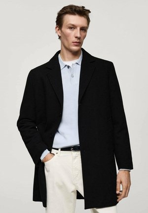 Cappotto di lana nera con design su misura e chiusura a un bottone, indossato sopra una polo azzurro chiaro e pantaloni bianchi.