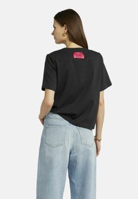 Schwarzes T-Shirt mit kurzen Ärmeln und einem pinken Etikett auf der Rückseite; hat einen lässigen Schnitt und weichen Stoff. Kombiniert mit hellblauen hochgeschnittenen Jeans.