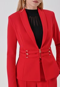 Rode op maat gemaakte blazer met een getailleerde taille, voorzien van gouden hardware en een ritssluiting. Glad materiaal en gestructureerde revers.