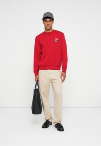 Sweatshirt rouge avec logo sur la poitrine, pantalon beige, tenant un sac fourre-tout noir, portant une casquette grise et des chaussures noires.