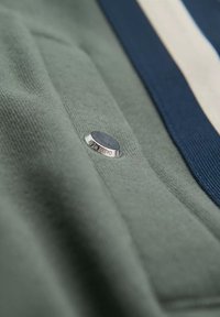 Grüner Stoff mit glatter Textur zeigt ein silbernes, rundes Metallelement mit der Aufschrift "Ombre Wear" und einem navyblauen Streifendetail.